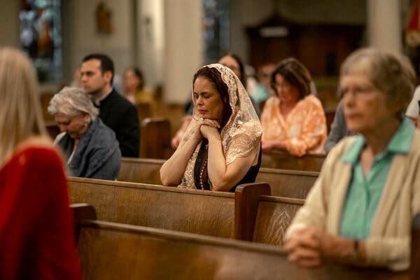 Several people sit in wooden pews, including some with eyes closed or heads bowed. 