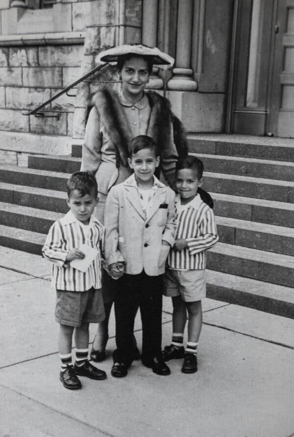A black and white image of a woman in a fur shawl and wide hat, standing behind three young boys holding hands, in front of concrete church steps. 