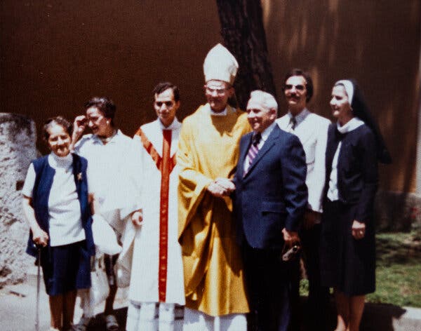 In a grainy photograph, people stand together in a group smiling. Some are wearing robes.